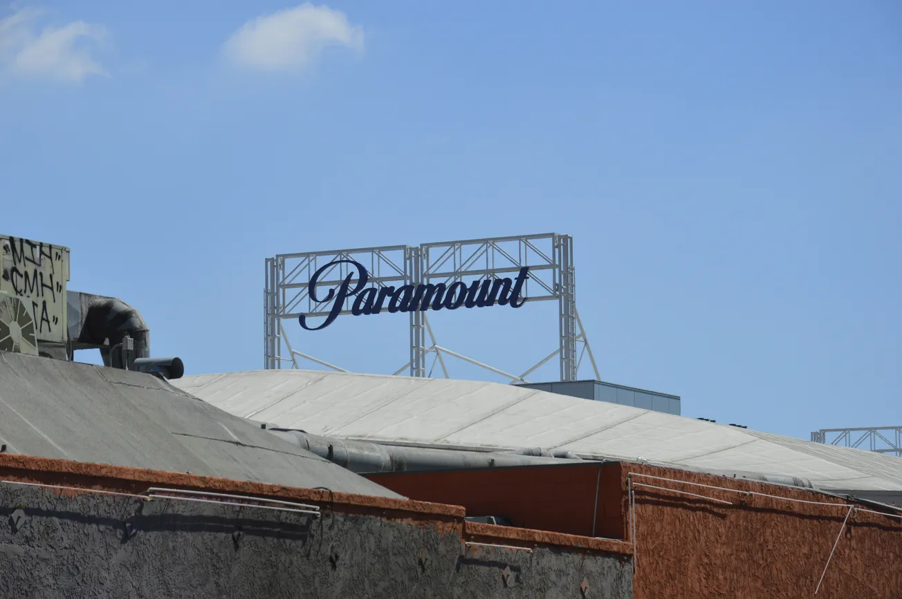 A large "Paramount" sign in blue script sits atop a building with an industrial rooftop. The sky is clear with a few clouds, conveying a sunny day.