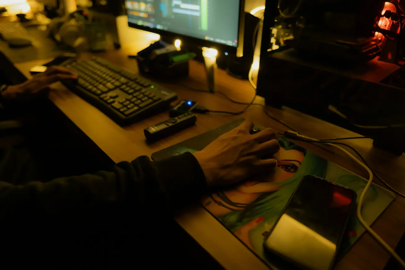 A person sitting at a desk in a dimly lit room, using a computer mouse. A glowing screen, keyboard, and phone nearby create a focused, tech-centric atmosphere.