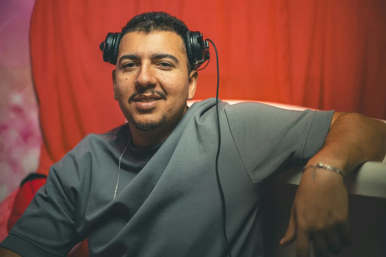 Smiling man wearing headphones rests his arm on a bar, set against a soft red backdrop. Relaxed ambiance with casual attire and dim lighting.