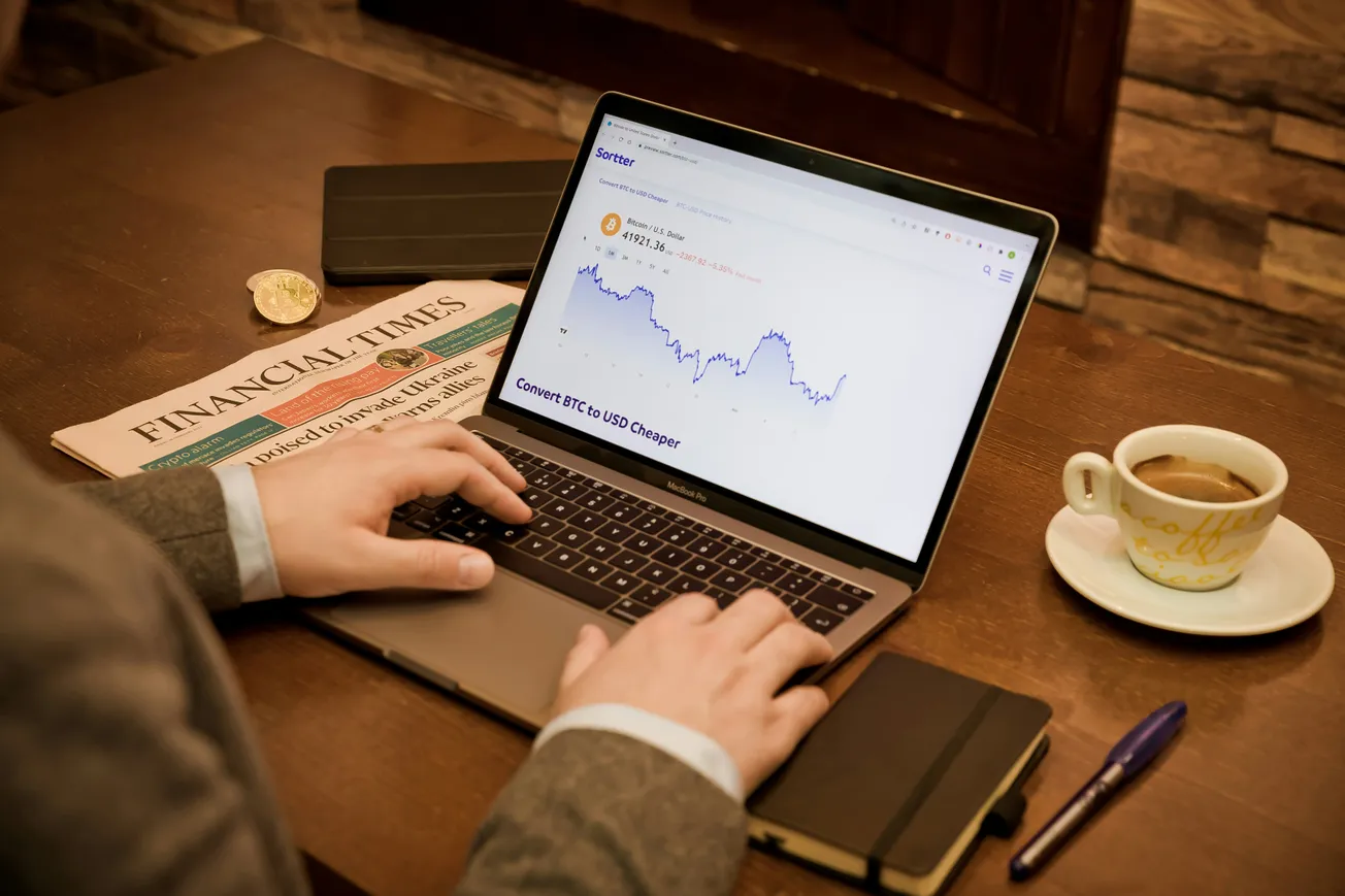 A person in a suit works on a laptop displaying a cryptocurrency chart. Nearby are a Financial Times newspaper, a notebook, a pen, and a cup of coffee on a wooden table.