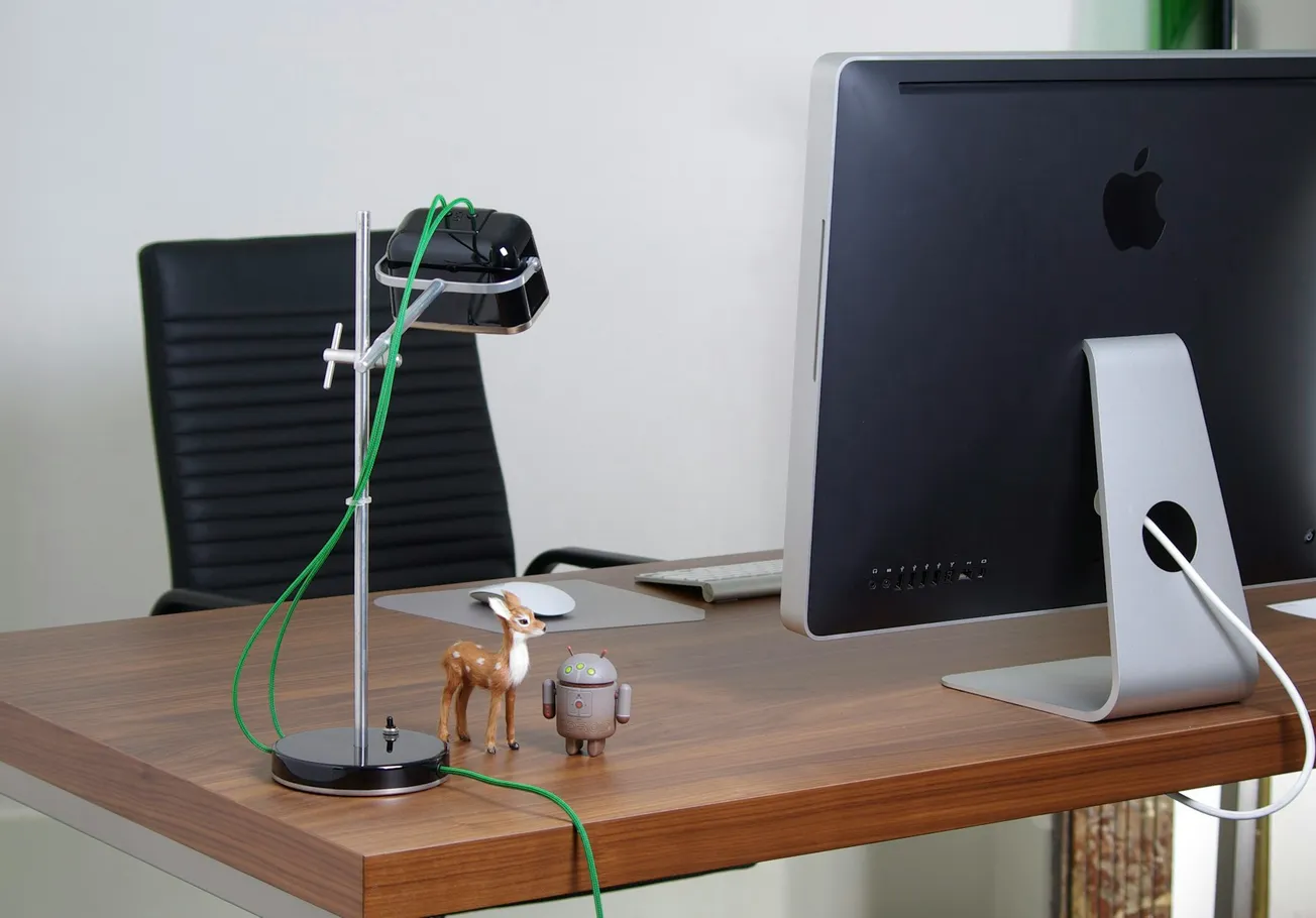 A modern office desk with a large computer monitor, black swivel chair, desk lamp with a green cable, and small toy figures, conveying a tidy workspace.