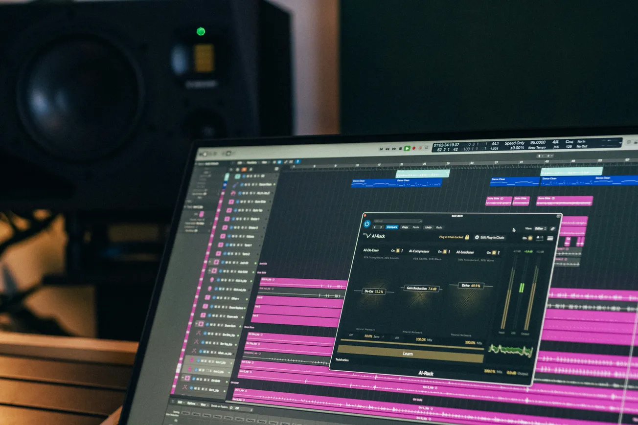 A computer screen displaying audio editing software with various tracks in pink and blue. A large speaker is visible in the background. The scene conveys a focused, creative atmosphere.