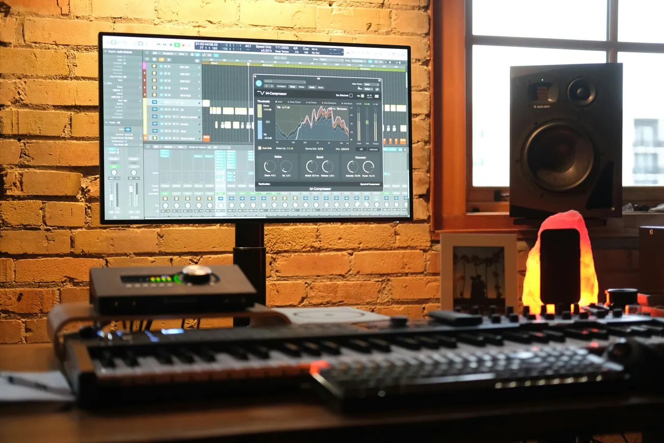 Music studio setup with a monitor displaying audio mixing software, keyboards, and speakers, set against a brick wall. Warm lighting creates a cozy ambiance.