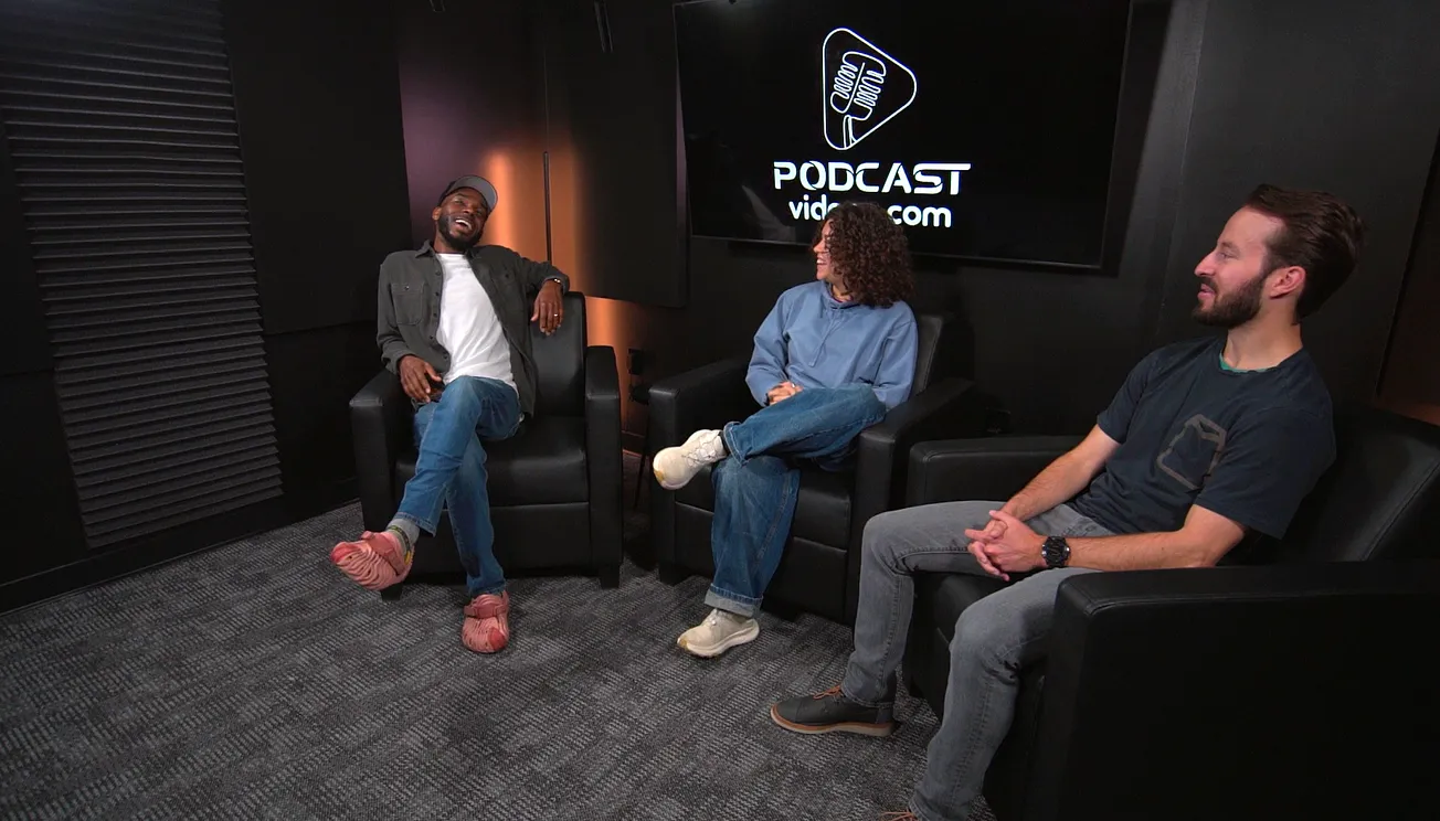 Three people sit in a dark podcast studio, smiling and engaged in conversation. The backdrop features a large screen with a podcast logo.