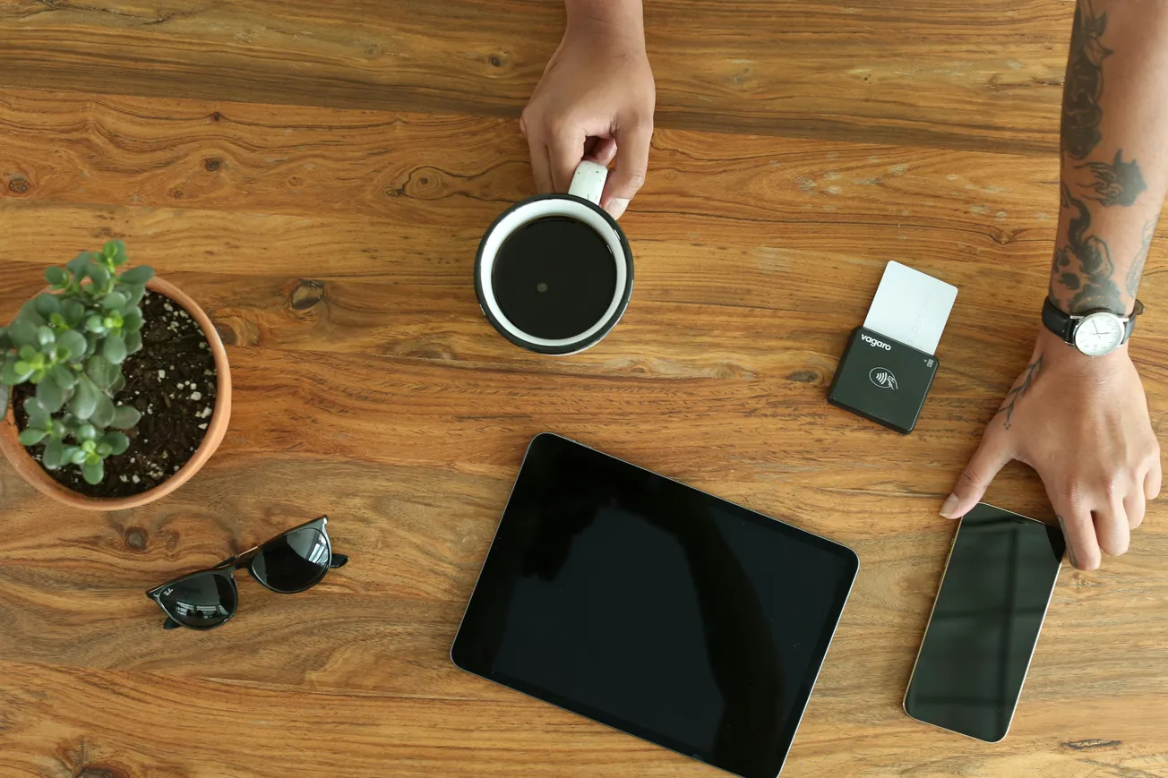 A wooden table with a hand holding a coffee cup, a tablet, smartphone, sunglasses, potted plant, and a payment terminal. Modern, relaxed workspace vibe.