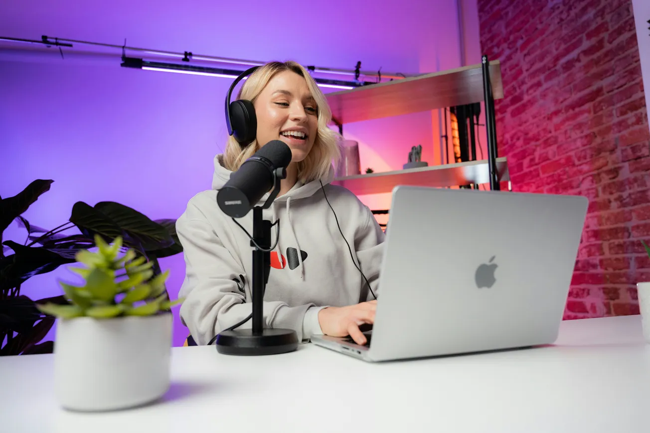 Smiling woman wearing headphones records a podcast at a laptop with a microphone. She's seated at a white desk with plants, against a purple-lit background.