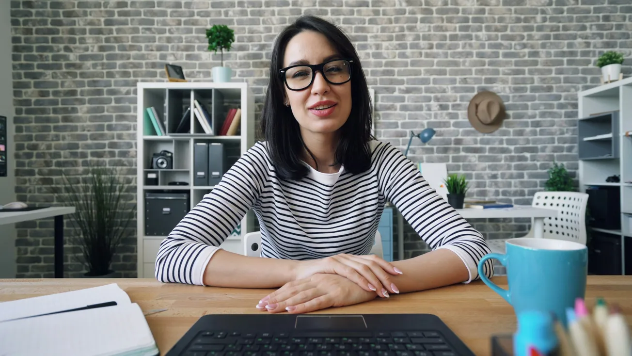 A woman with glasses and a striped shirt sits at a desk, smiling. Behind her is a gray brick wall, and a shelf with books and plants, creating a cozy office vibe.
