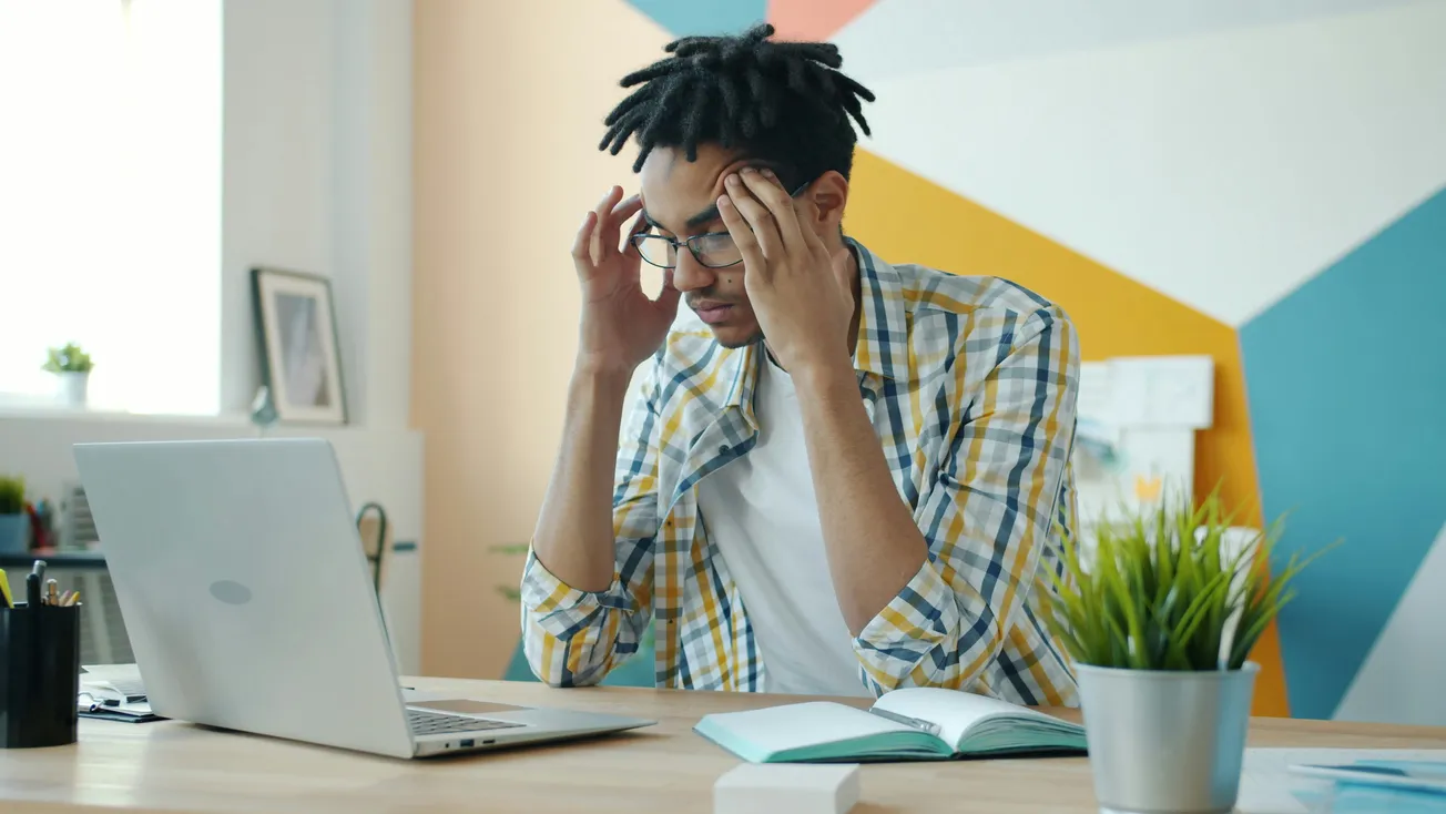 A man sits at a desk in a colorful office, holding his head and looking frustrated. An open laptop, notebook, and potted plant are on the desk.
