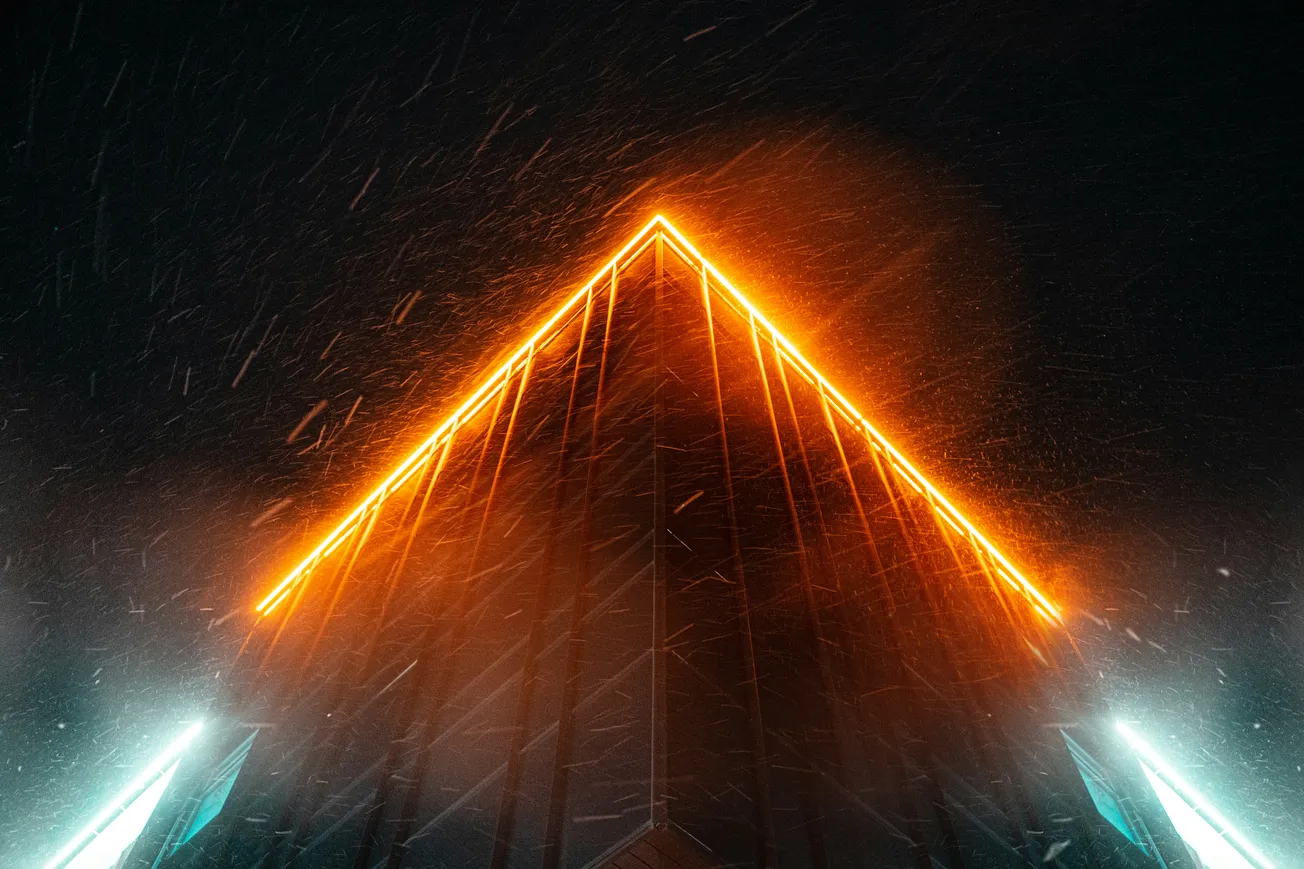 A triangular building facade illuminated with bright orange neon lights against a dark, snowy night sky. The scene evokes a futuristic and dynamic mood.