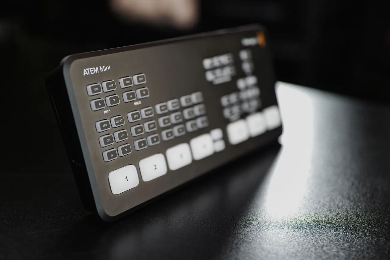 A black ATEM Mini video switcher with numerous buttons sits on a reflective dark surface, capturing a sleek and modern tech aesthetic.