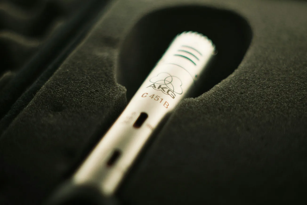 Close-up of a silver microphone labeled "AKG C 451 B" nestled in a dark foam case, conveying a sense of professional audio equipment ready for use.