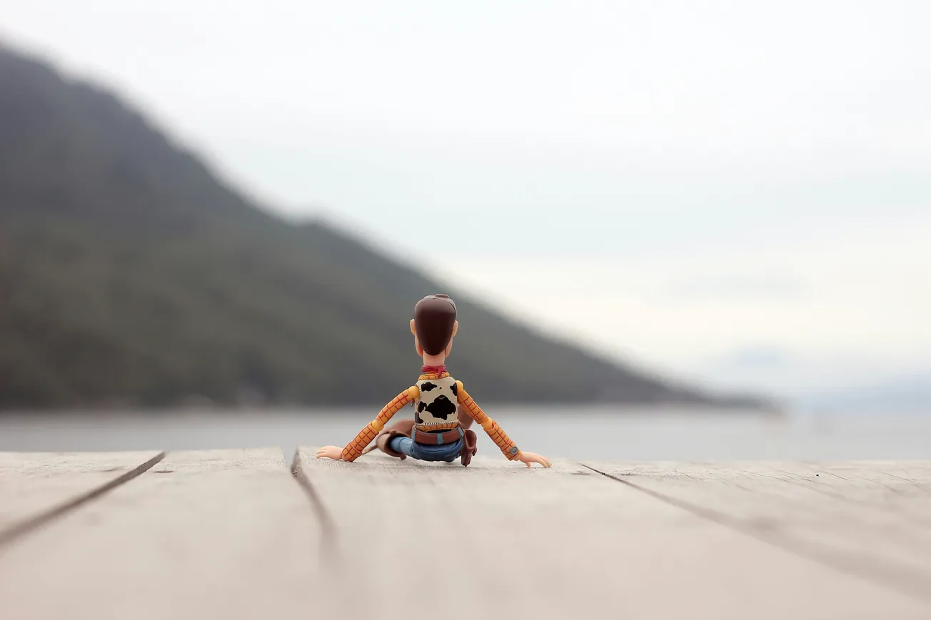Toy figure in cowboy attire sits on a wooden surface, gazing out at a misty, mountainous landscape beside a serene body of water, evoking nostalgia.