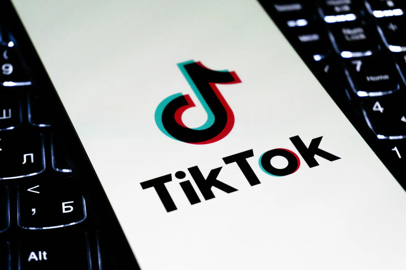 Smartphone screen displays the TikTok logo, a musical note icon, over a white background. The device rests on a lit keyboard, symbolizing technology.