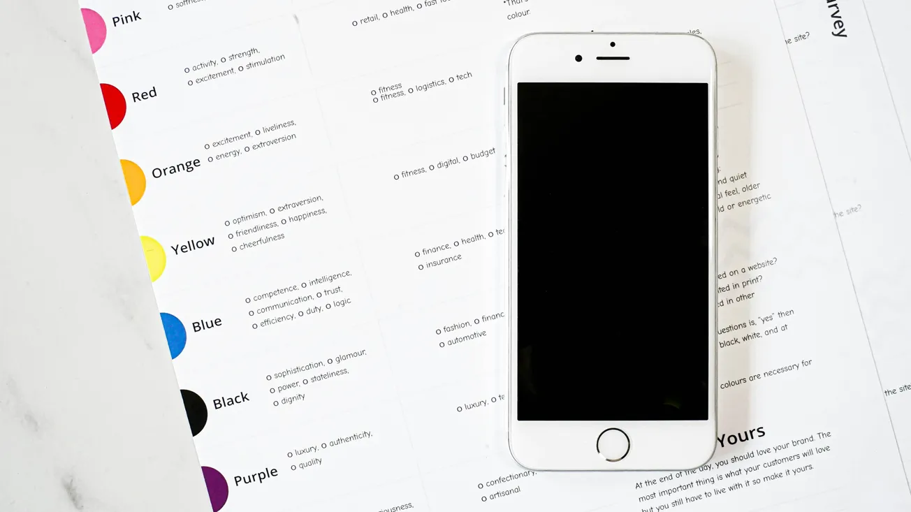 A smartphone with a blank screen is placed on a spread of printed pages featuring color swatches and descriptions. The setting is bright and minimalistic.