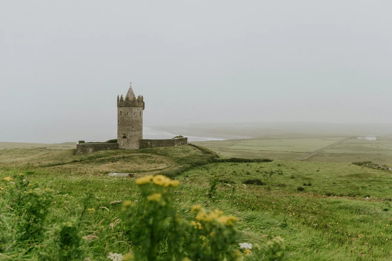 A solitary stone castle tower stands amid lush green fields under a hazy sky. Yellow wildflowers in the foreground add a touch of brightness to the misty scene.