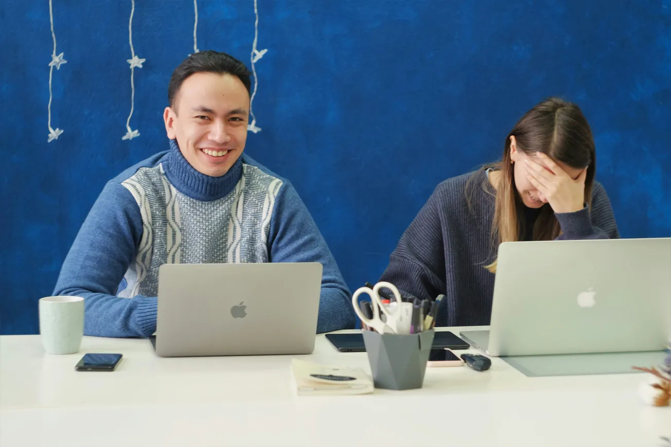 Two people sit at a desk with laptops and office supplies. The man smiles warmly, while the woman covers her face, conveying a lighthearted tone.