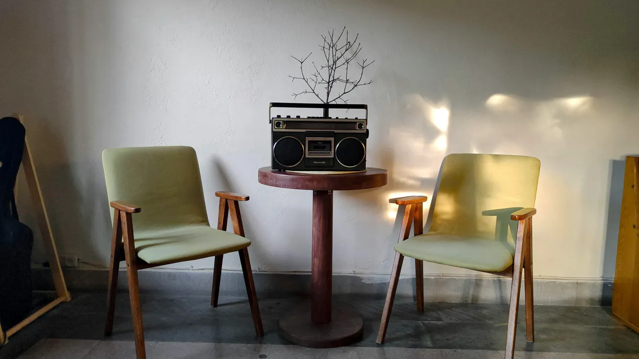 Two mid-century modern chairs flank a round wooden table with a vintage radio and a small tree branch arrangement, set against a softly lit wall.