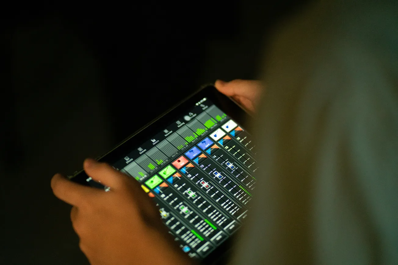 Hands holding a tablet displaying a colorful digital audio mixer interface with sliders and levels. The ambient lighting is dim, creating a focused mood.