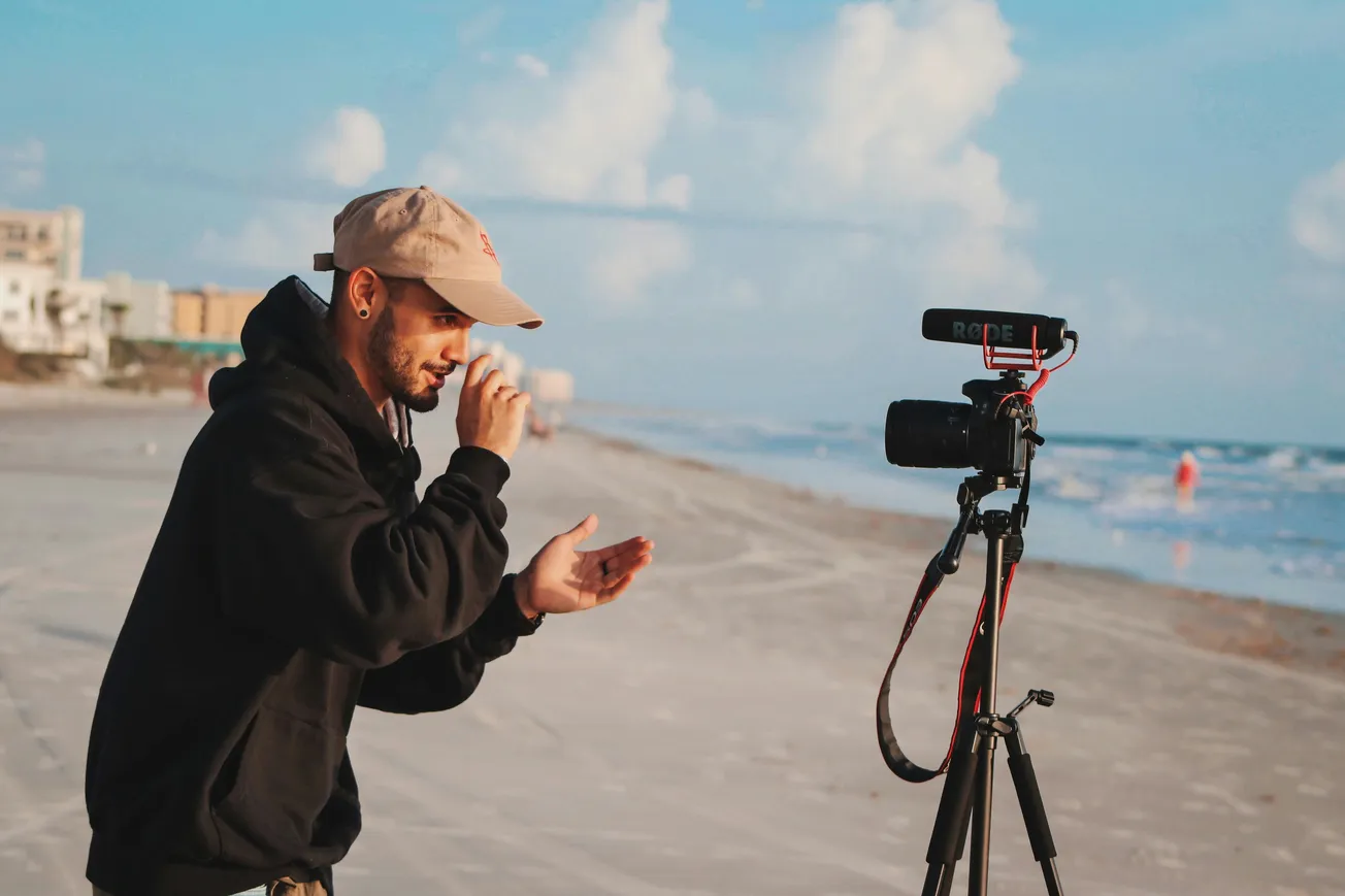 A man in a hoodie and cap gestures animatedly by a camera on a tripod, filming on a sandy beach under a bright, cloudy sky.