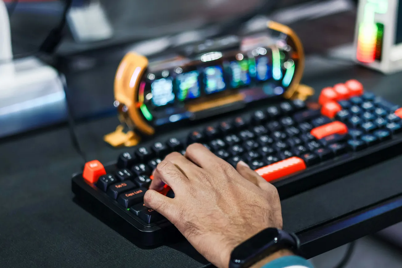 A hand rests on a colorful gaming keyboard with vibrant keys. Behind it, a futuristic, illuminated device displays various data. The scene conveys a tech-savvy, engaged atmosphere.