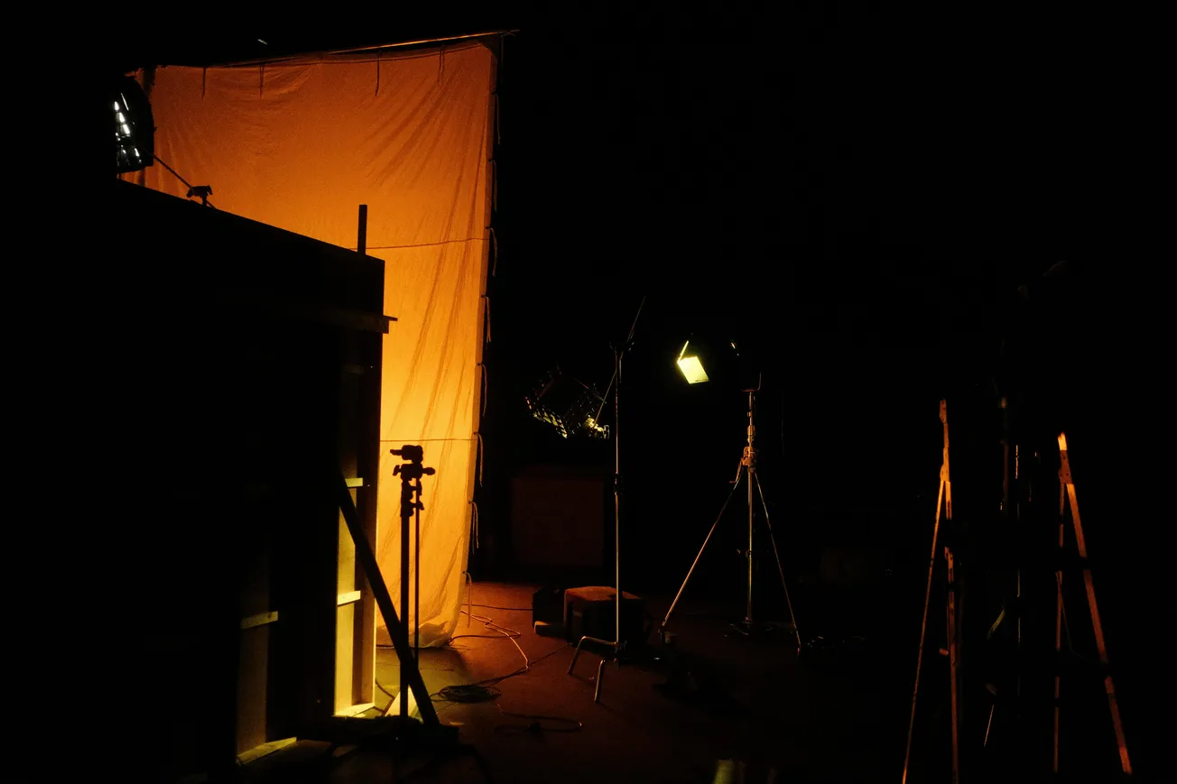Dimly lit room with a glowing orange backdrop and various light stands. The scene conveys a warm, moody atmosphere, suggesting a film or photo set.