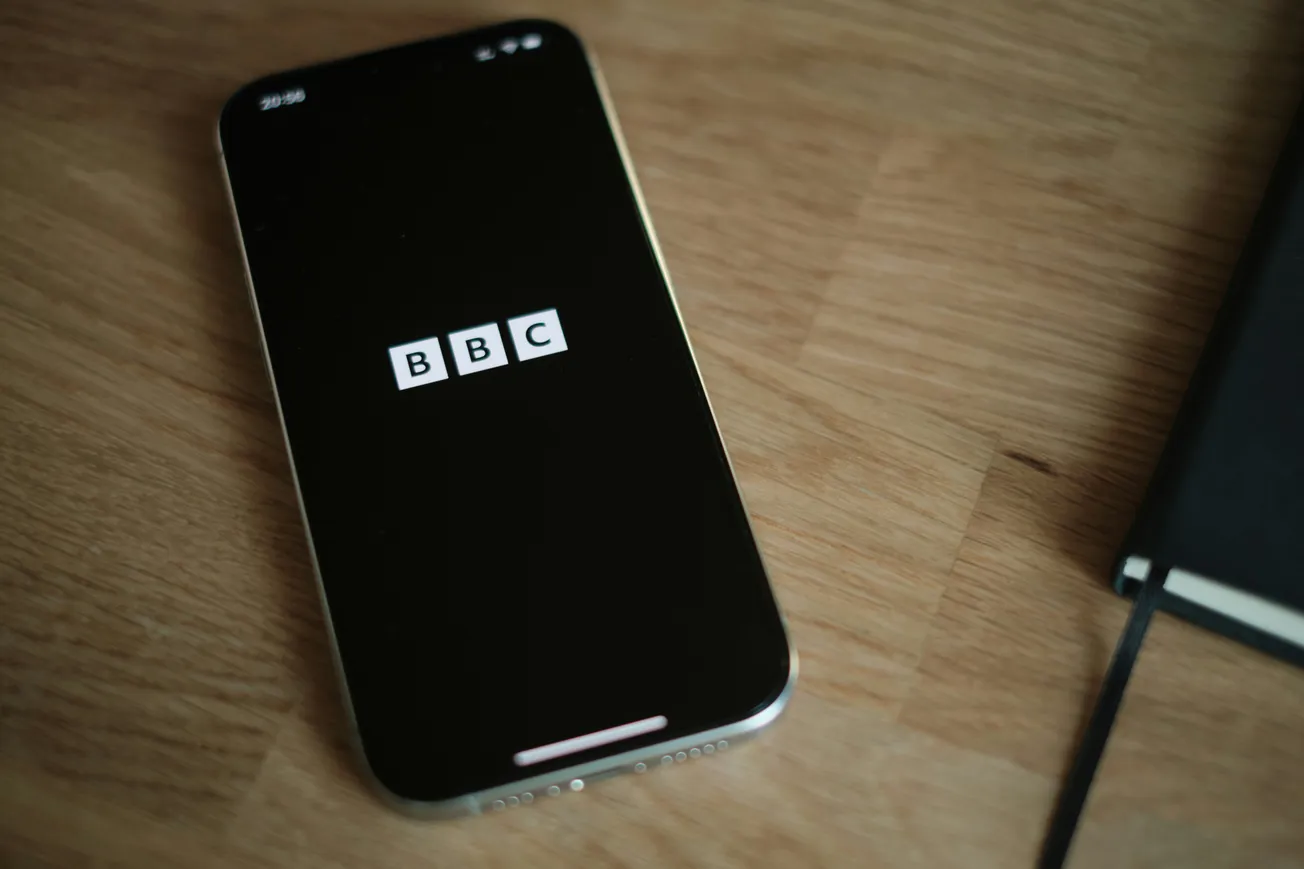 Smartphone on a wooden table displaying the BBC logo on screen, next to a closed black notebook. The scene feels modern and professional.