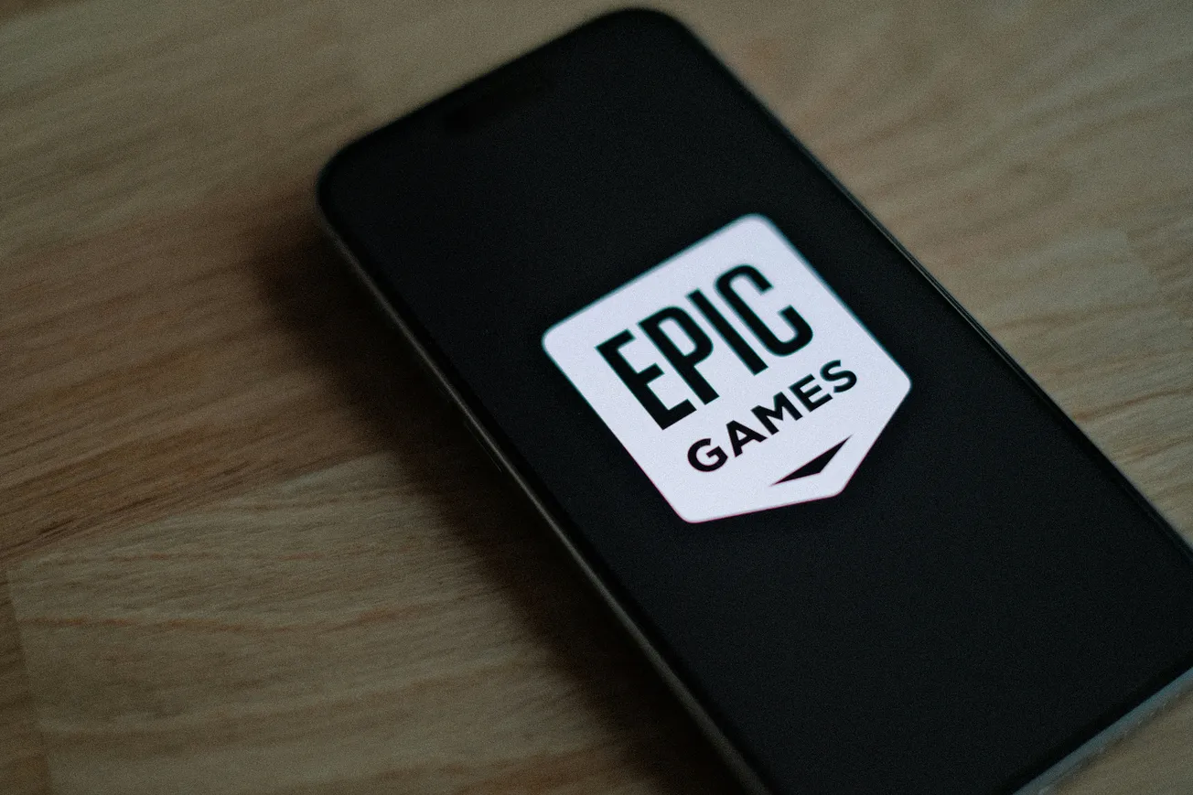 A smartphone on a wooden surface displays the Epic Games logo, its white icon and bold text glowing against the dark screen, evoking a modern gaming vibe.