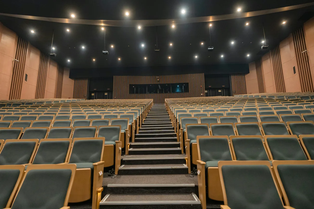 An empty auditorium with rows of gray upholstered seats, wooden panels on the walls, and ceiling lights illuminating the spacious, quiet interior.