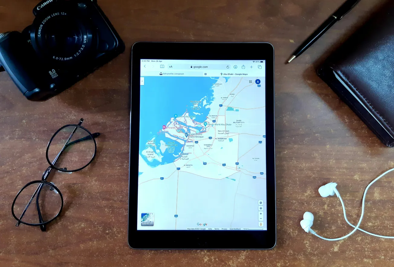 Tablet displaying a coastal map on a wooden desk, surrounded by a camera, glasses, pen, leather notebook, and white earphones, suggesting travel planning.