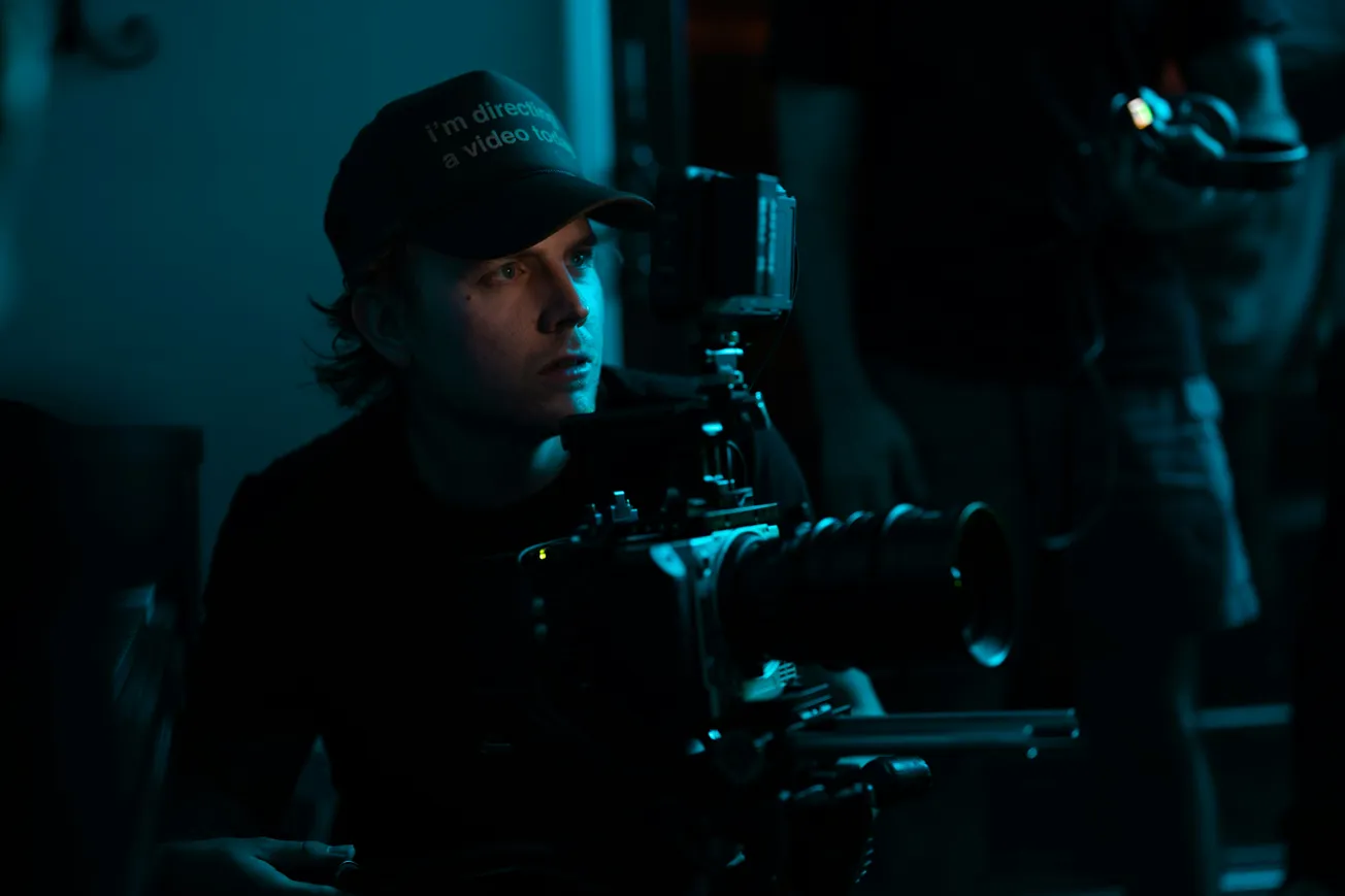 A person wearing a cap operates a camera, bathed in dramatic blue lighting. Focused expression suggests concentration and creativity in a film set environment.