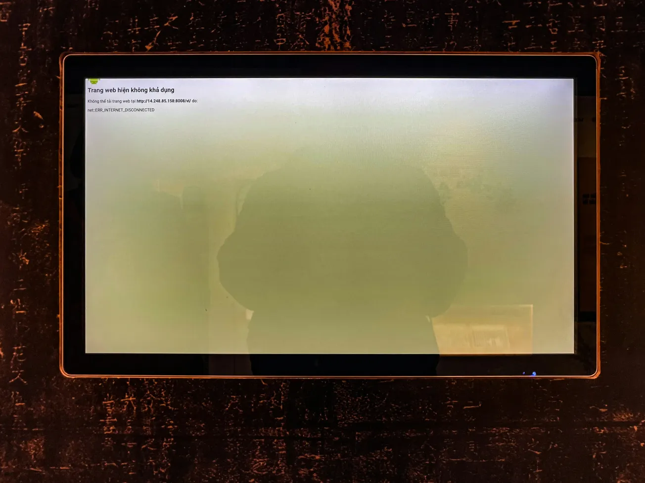 A touch screen displays an error message in Vietnamese on a black, textured background. The reflection of a person is faintly visible on the screen.