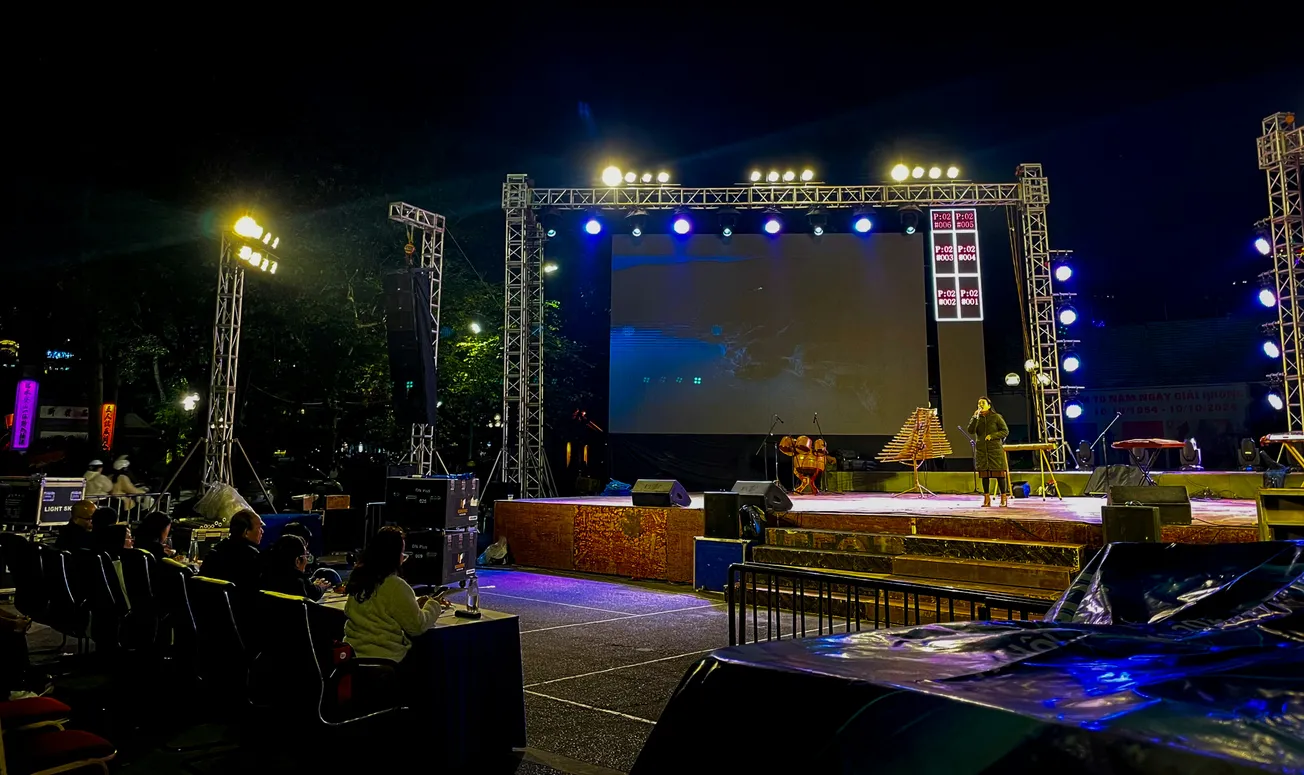 Outdoor stage at night with bright lights and large screen, hosting a performance. Small audience seated in the foreground. Energetic atmosphere.