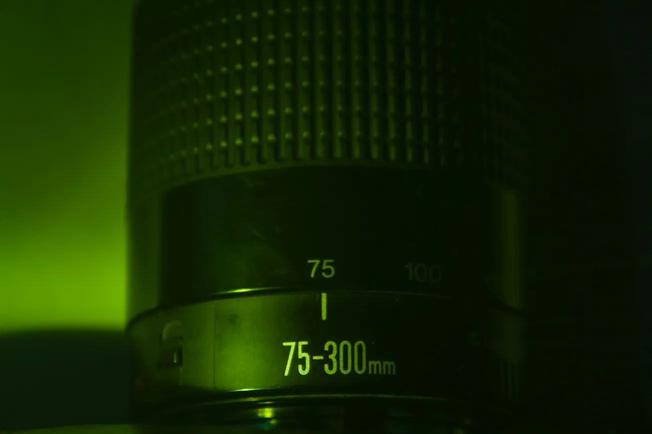 Close-up of a camera lens, labeled 75-300mm, in dark lighting with a green tint. The focus ring and textured grip are visible, evoking a moody tone.