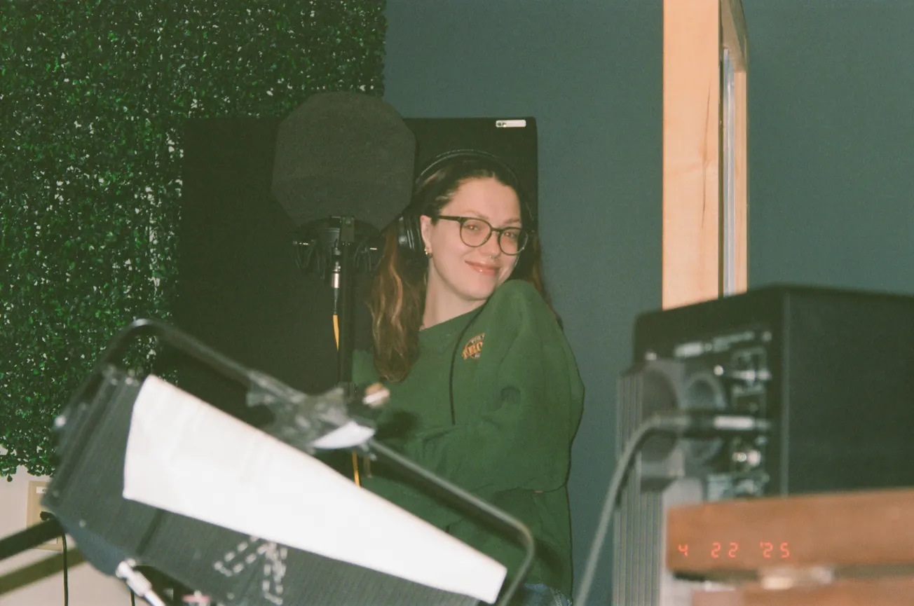 A person with glasses and a green sweatshirt smiles near a microphone in a cozy recording studio. The atmosphere feels relaxed and creative.
