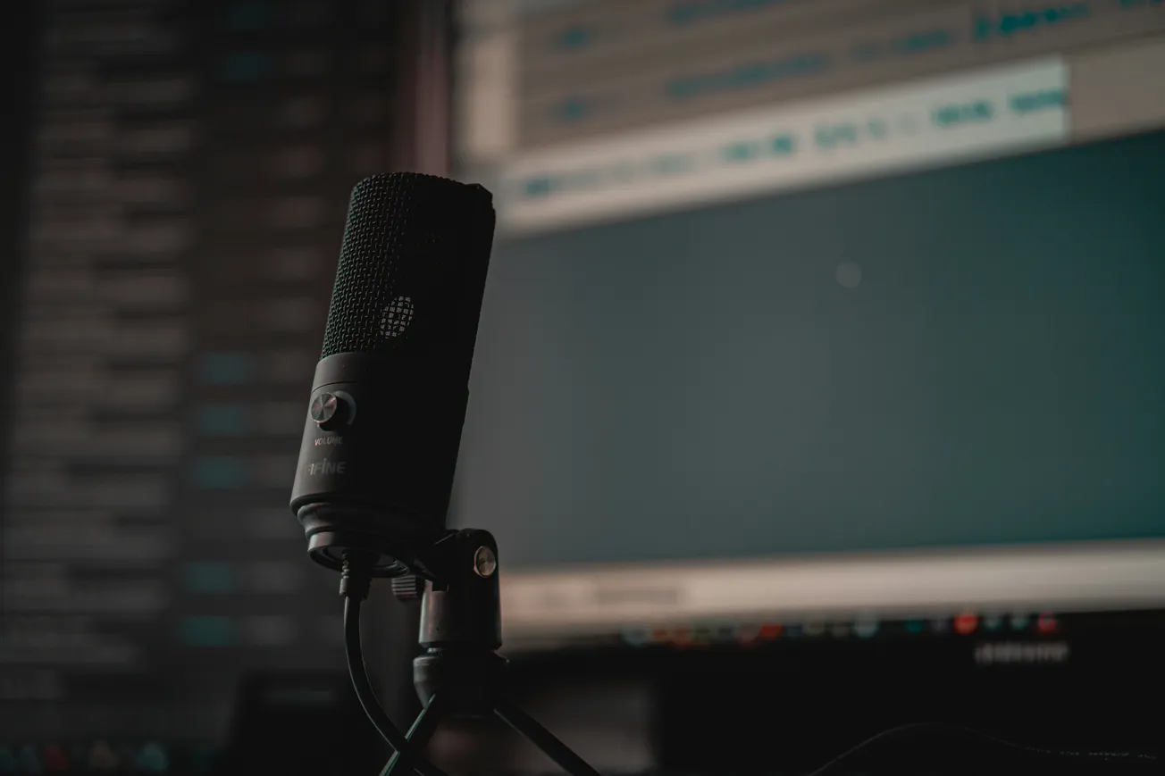 A black microphone is in focus against a blurred computer screen displaying audio editing software, creating a professional recording studio vibe.