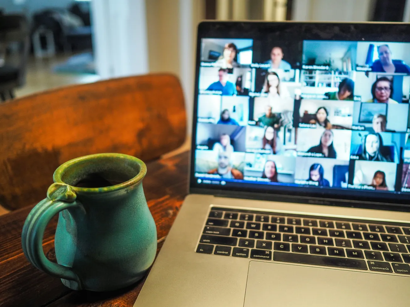 A laptop on a wooden table shows a video call with many participants. A green ceramic mug sits beside it, creating a cozy, work-from-home vibe.