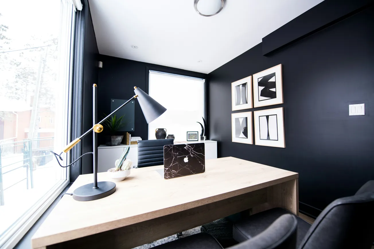 Modern office with a wooden desk, black lamp, and a laptop. Black walls display abstract art. Large window provides bright, natural light.
