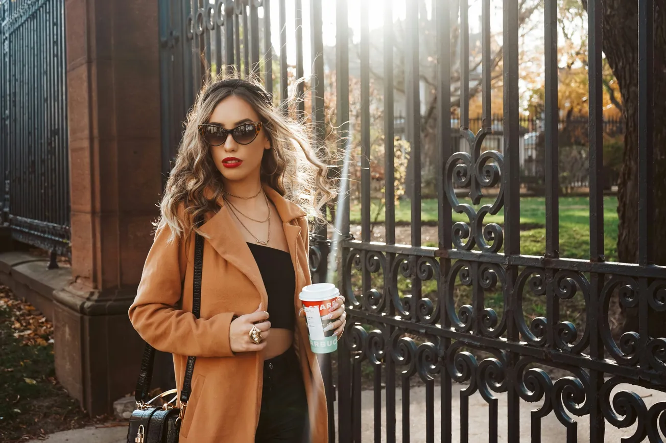 A woman wearing sunglasses and a coat holds a coffee cup, standing outdoors with a relaxed expression.
