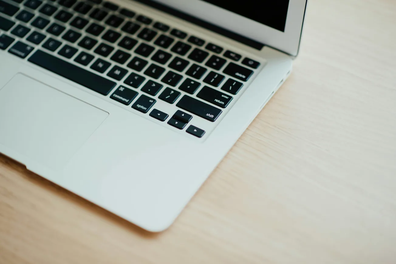 Close-up of an open laptop on a light wooden desk. The silver laptop shows part of the keyboard and touchpad, conveying a clean, modern workspace.