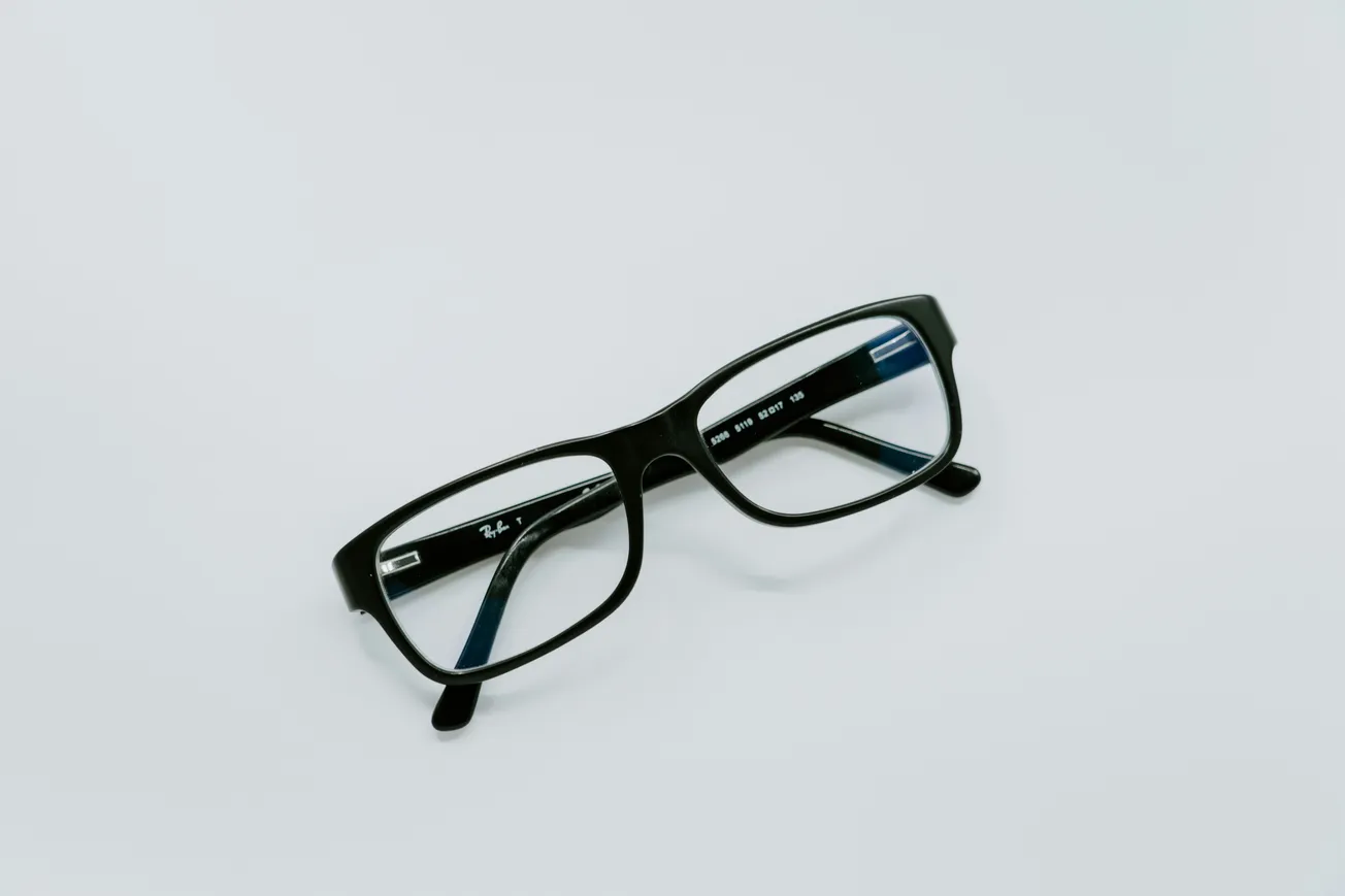 Black rectangular eyeglasses with a glossy frame lie on a white surface. The simple design conveys functionality and classic style.