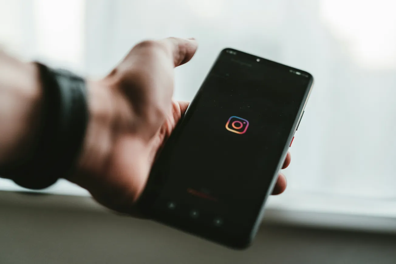 A person holds a smartphone displaying the Instagram app icon, with the phone in focus and a blurred background. The mood is casual and tech-oriented.