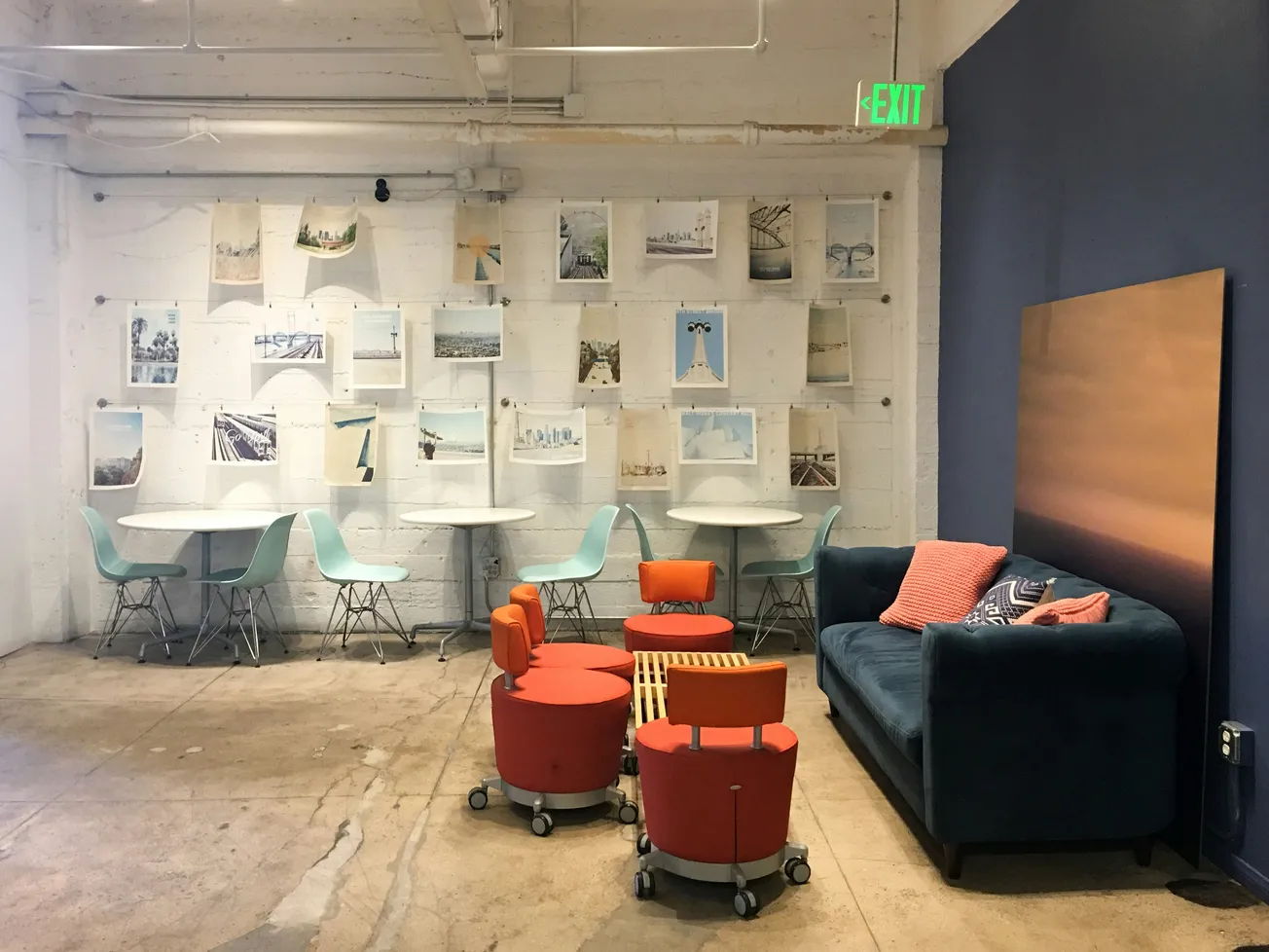 Cozy lounge area with a blue sofa, orange cushions, and rolling stools on a concrete floor. Art-covered white wall and mint chairs create a modern vibe.