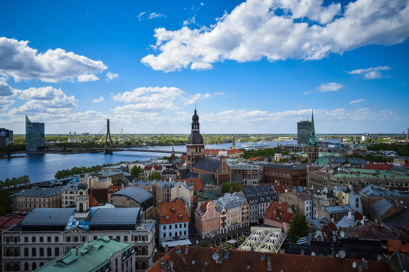 Aerial view of Riga cityscape with a backdrop of a bright blue sky and fluffy clouds. Historic buildings, a river, and modern bridges are visible. Vibrant and scenic.