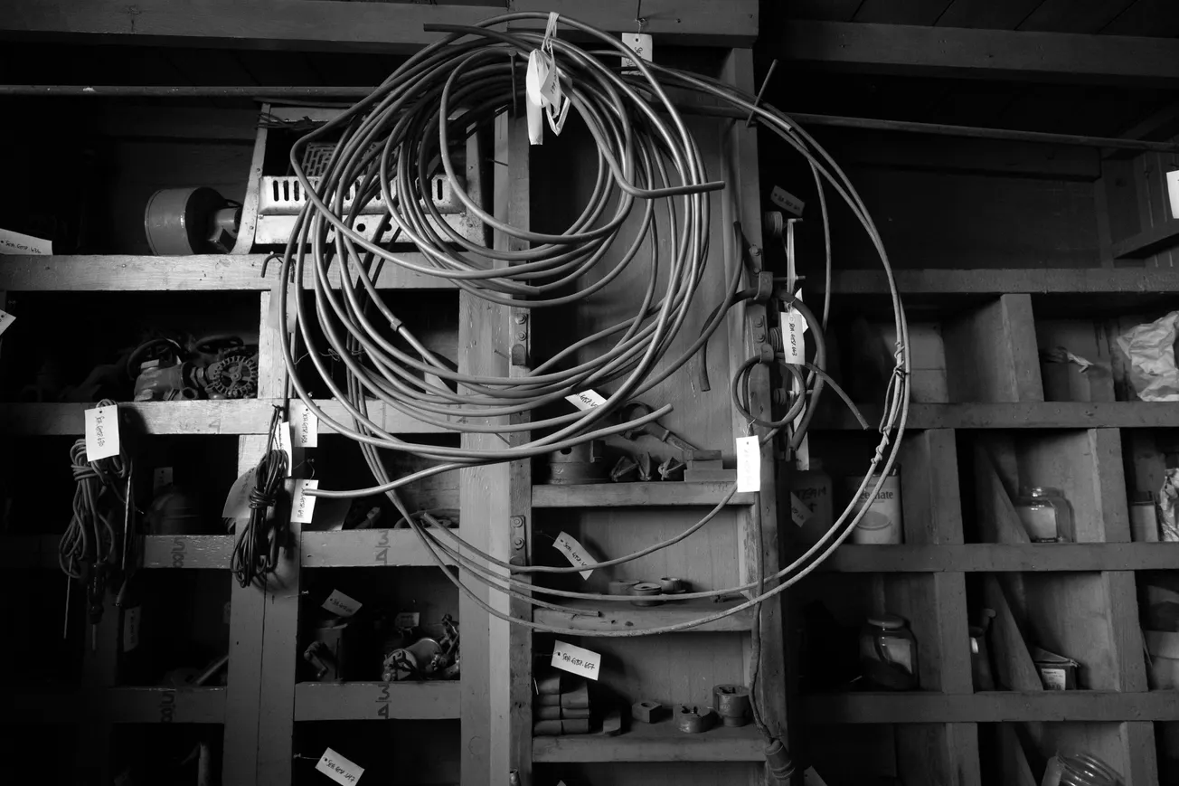 A black and white image of a cluttered workshop wall with neatly coiled cables and wires hanging. Numerous labeled shelves hold various tools and items.