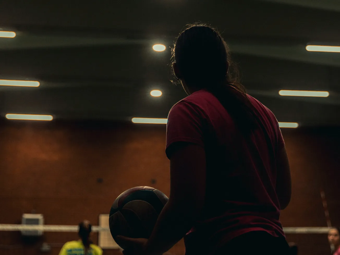 Silhouetted person holding a volleyball indoors, facing a dimly lit court. Overhead lights create a dramatic, focused atmosphere.