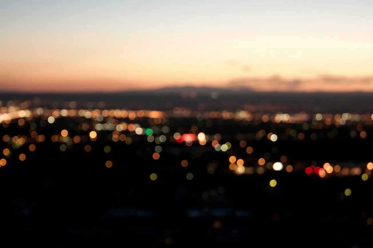Blurry cityscape at twilight with out-of-focus lights glowing in warm tones against a gradient sky transitioning from soft orange to deep blue. Calm and serene atmosphere.