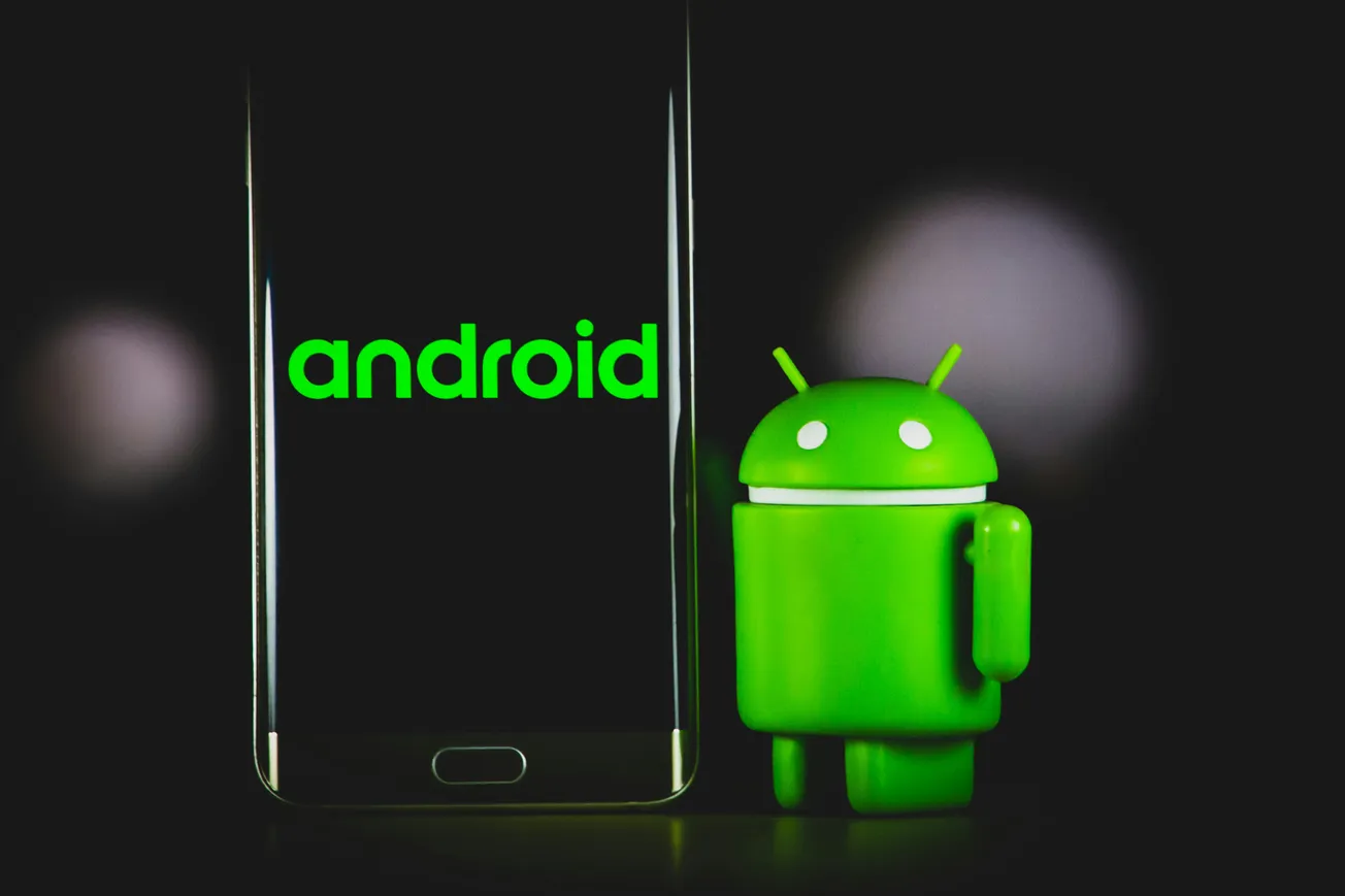 A smartphone displaying the word "android" in bright green sits next to a small, green Android robot figure. The background is dark, creating a tech-focused tone.