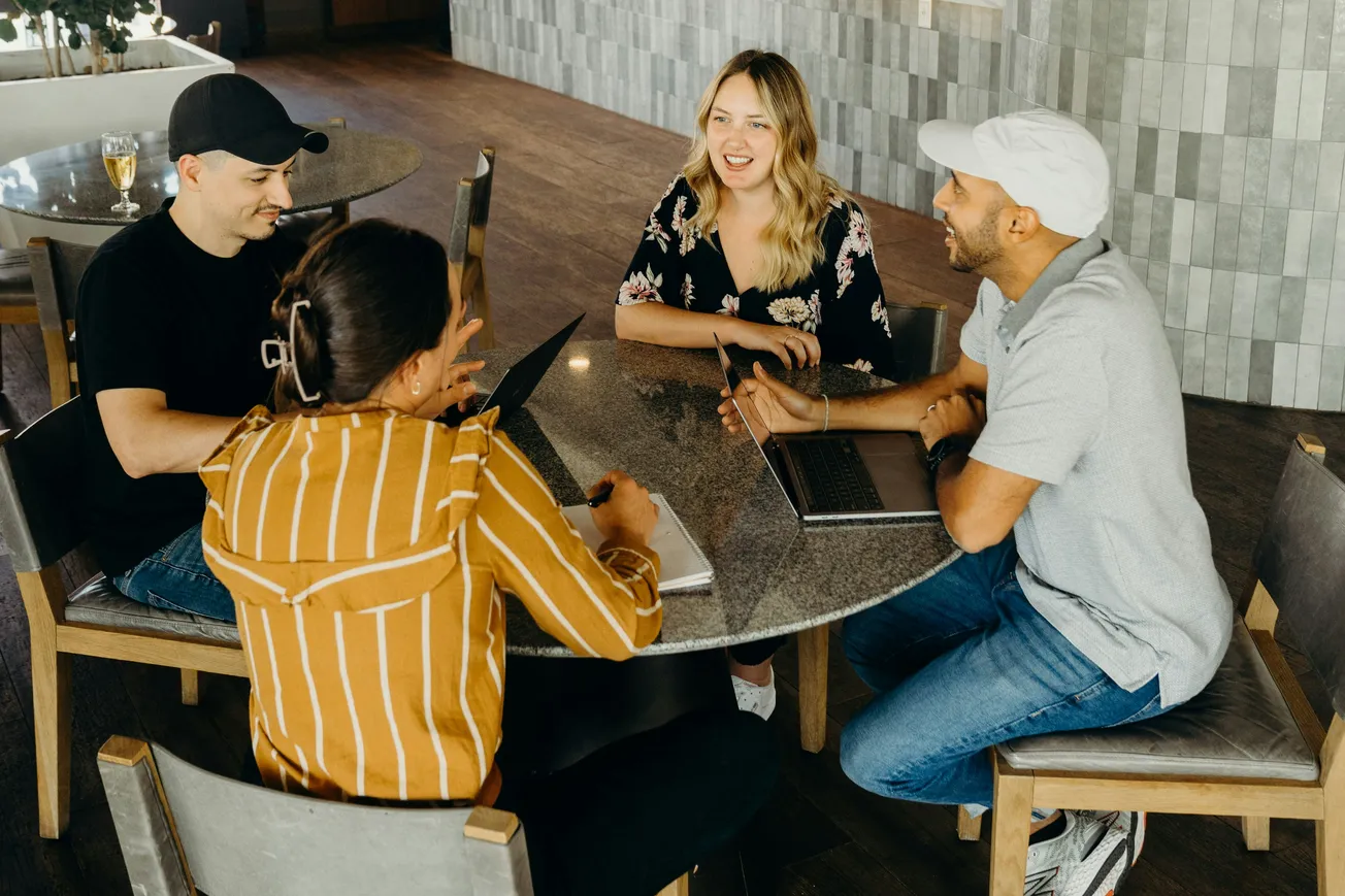 Four people sitting together at a table with their laptops, engaged in conversation and laughter.
