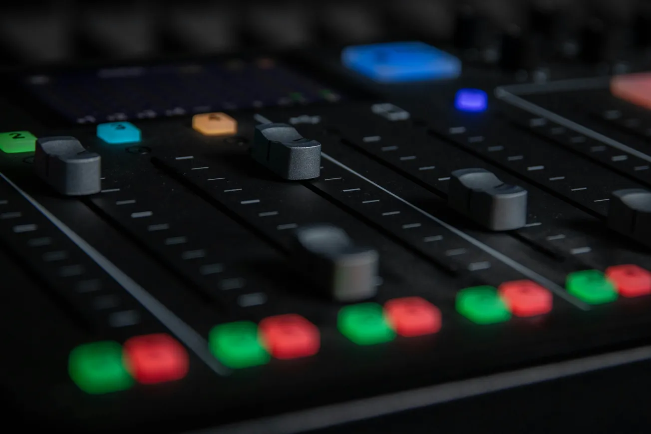 Close-up of a sound mixing board with sliders and colorful buttons. Dim lighting gives a focused, professional studio atmosphere.