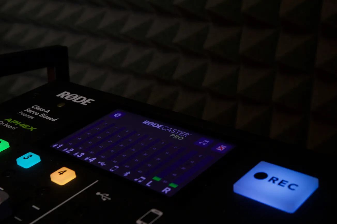 Close-up of a RØDECaster Pro mixing console in dim light, highlighting the glowing multicolored buttons and blue REC button, creating an ambient studio vibe.